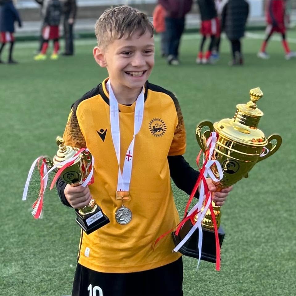 Sandro Abesadze smiling on the football pitch while holding two trophies and wearing a medal